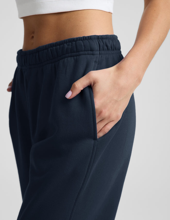 Woman wearing a white ribbed tank top and blue joggers against a plain background.