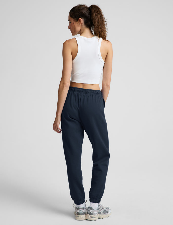 Woman wearing a white ribbed tank top and blue joggers against a plain background.