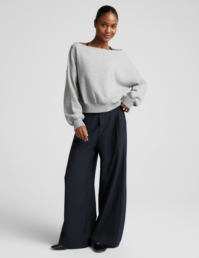 A woman wearing a light gray off-the-shoulder knit sweater and dark navy pants, photographed against a plain light gray studio background.