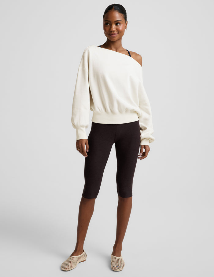 Woman wearing a white off the shoulder fleece sweatshirt and brown leggings against a plain background.