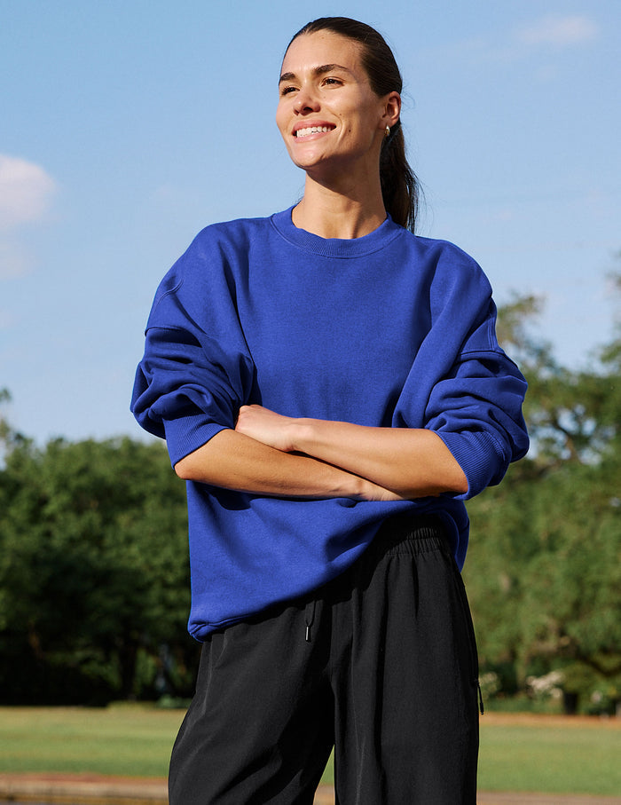 A woman wearing a vibrant royal blue crewneck sweatshirt and black sweatpants, standing outdoors with her arms crossed under a clear blue sky.