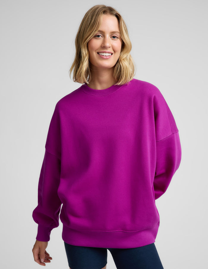 Woman wearing a purple oversized crewneck sweatshirt and navy blue biker shorts against a plain background.