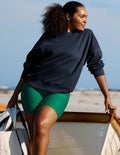 A woman wearing a dark navy crewneck sweatshirt and bright green biker shorts leans on the edge of a wooden boat at a beach.