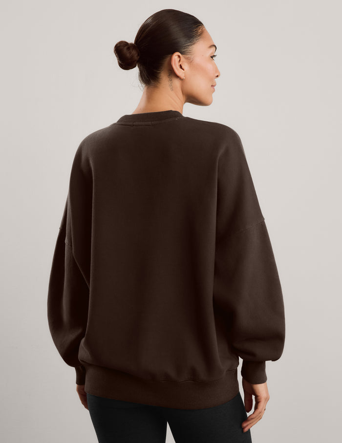 Woman wearing a brown oversized crewneck sweatshirt and black leggings against a plain background.
