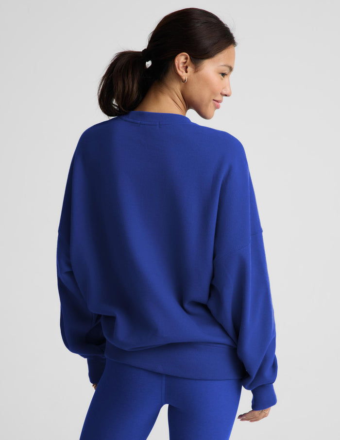 A woman shown from the back wearing a vibrant royal blue crewneck sweatshirt and matching blue leggings against a light gray studio background.