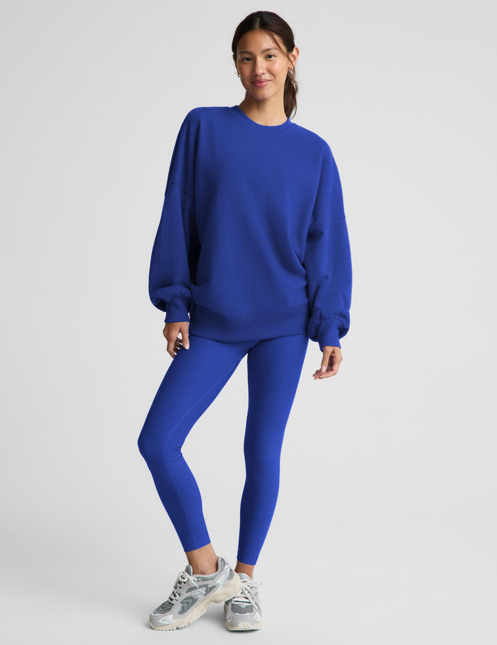 A woman modeling a matching royal blue sweatshirt and leggings set, paired with gray and white sneakers, against a light gray studio background.
