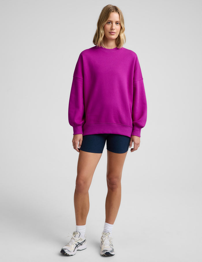 Woman wearing a purple oversized crewneck sweatshirt and navy blue biker shorts against a plain background.