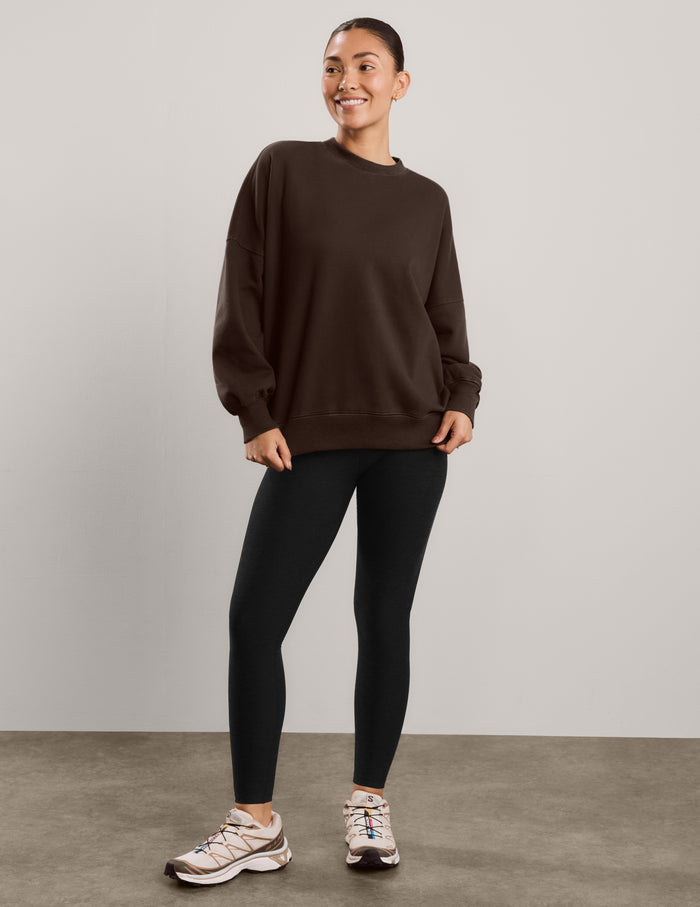 Woman wearing a brown oversized crewneck sweatshirt and black leggings against a plain background.