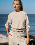 A woman wearing a light beige cropped sweatshirt and matching sweatpants stands outdoors near the water, with rocks and a blue sky in the background.