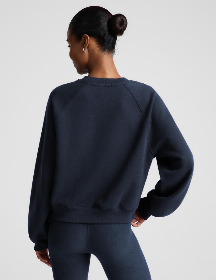 A woman wearing a dark navy blue crewneck sweatshirt and matching navy blue leggings, photographed against a solid light gray studio background.