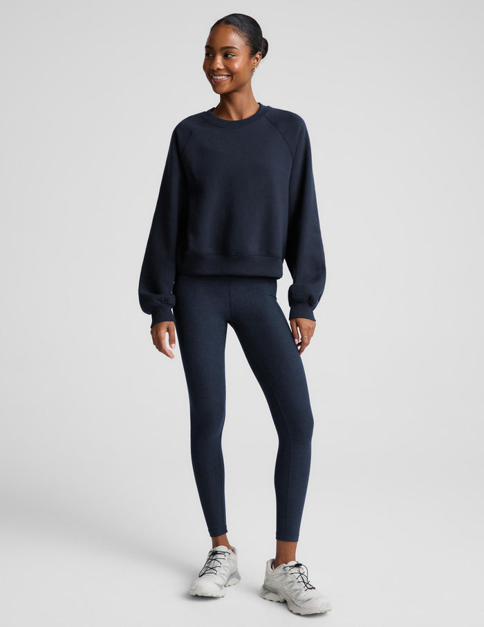 A woman wearing a dark navy blue crewneck sweatshirt and matching navy blue leggings, photographed against a solid light gray studio background.