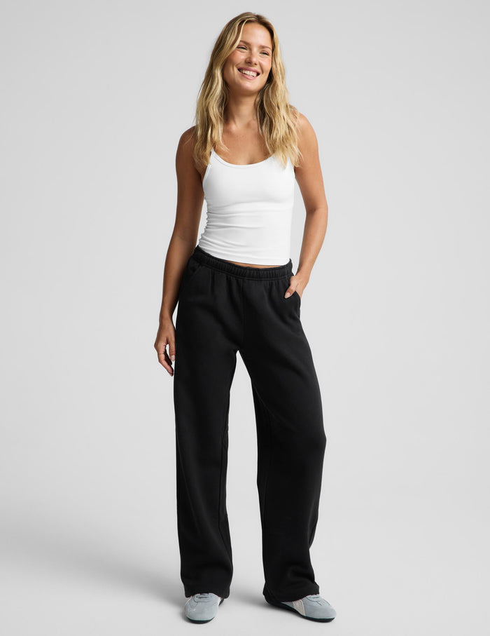 A woman modeling black wide-leg sweatpants with a white tank top and gray slip-on shoes, standing against a light gray studio background.