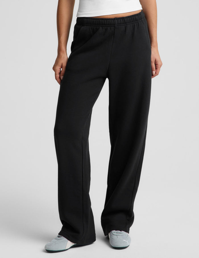 A woman modeling black wide-leg sweatpants with a white tank top and gray slip-on shoes, standing against a light gray studio background.