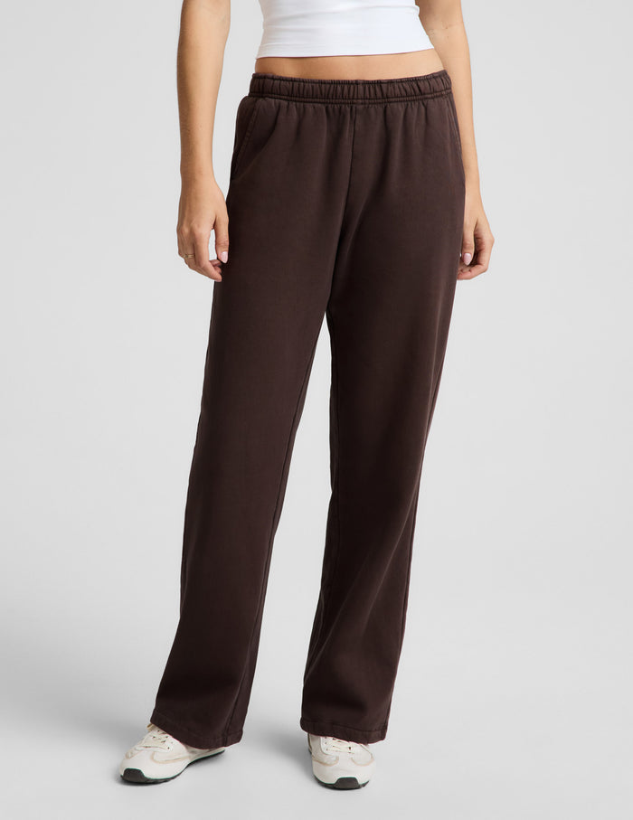 Woman wearing a white tank top and brown straight leg sweatpants against a plain background.