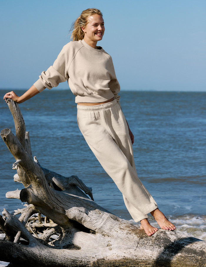A woman stands barefoot on driftwood at the beach wearing a light beige crewneck sweatshirt and matching sweatpants.