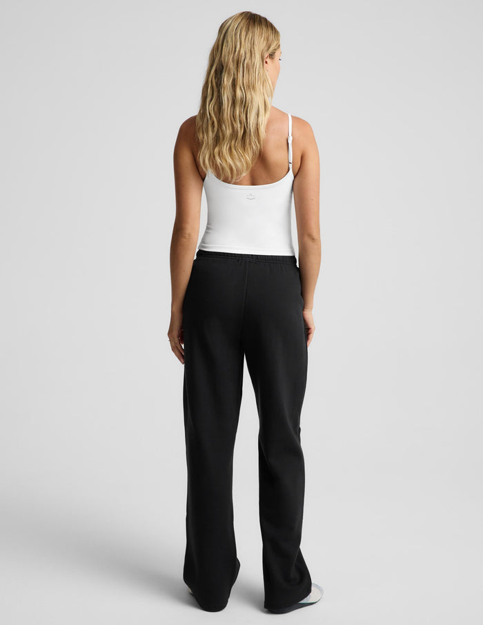 A woman modeling black wide-leg sweatpants with a white tank top and gray slip-on shoes, standing against a light gray studio background.