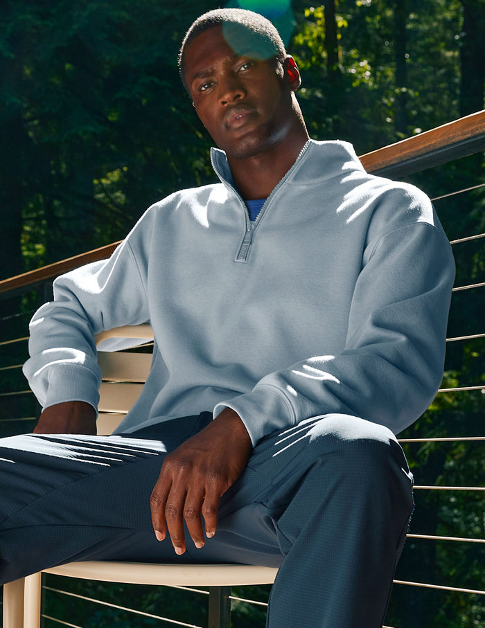 Man wearing a light blue pullover and dark pants sitting outdoors with greenery in the background.