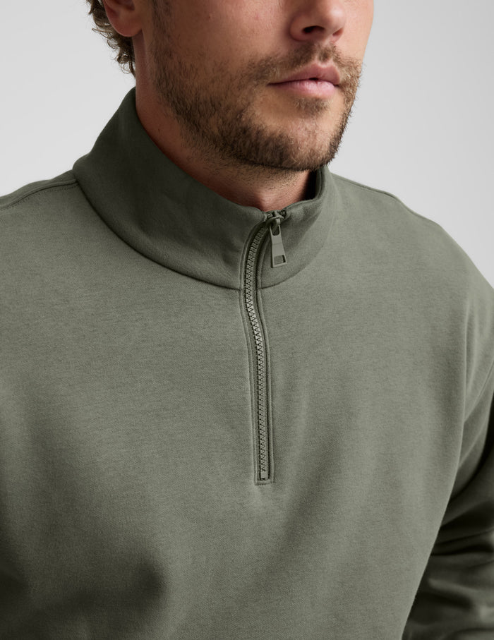 Man wearing a green quarter zip pullover and khaki pants with a plain background.