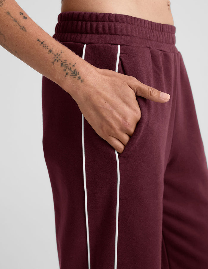 LuxeFleece Piper Wide Leg Pant in maroon burgundy with white side piping, elastic waist, and hand in the side pocket.