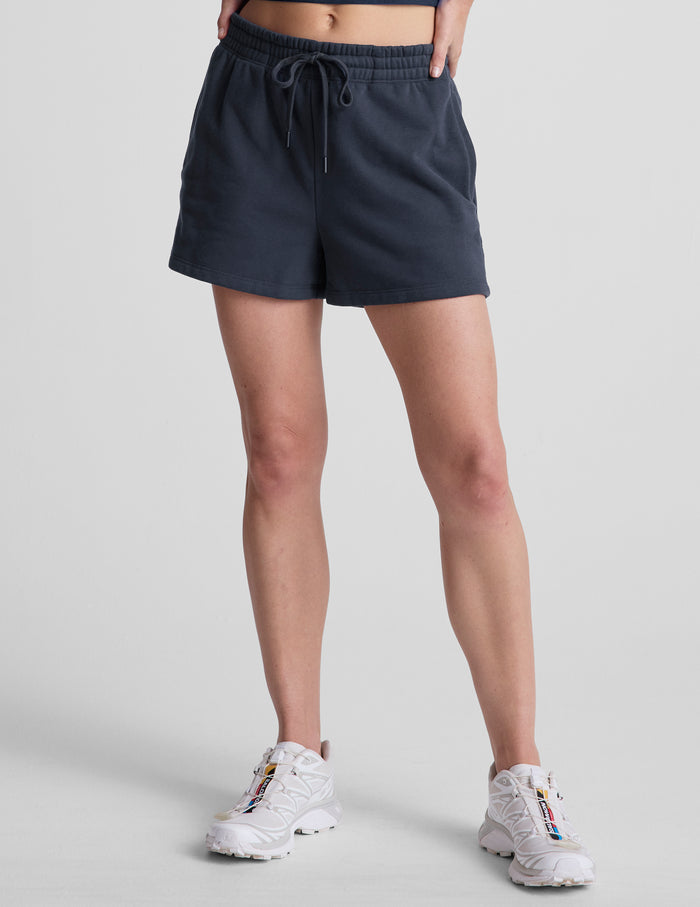 LuxeFleece Sweat Short in navy blue, featuring an elastic drawstring waistband and relaxed fit on a model in white sneakers.