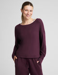 A woman modeling a matching deep plum purple loungewear set, featuring a long-sleeve boat-neck top and coordinating sweatpants, against a solid light gray background.