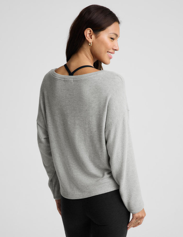 A woman shown from the back wearing a light gray knit sweater with a wide, slightly slouchy neckline and black leggings, photographed against a plain light gray studio background.