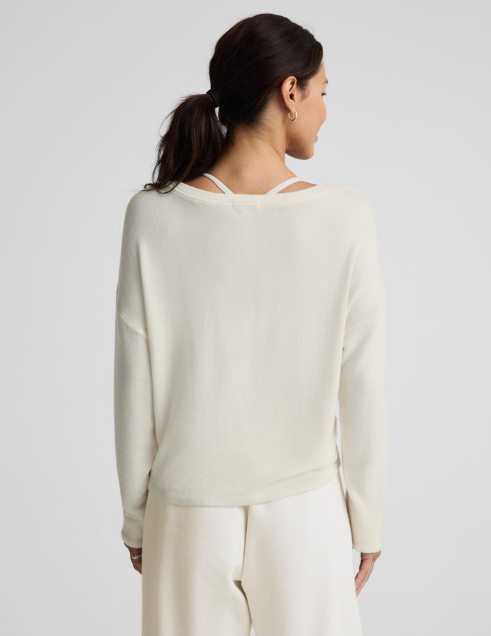 A woman shown from the back wearing a cream-colored knit sweater with a double-strap open neckline and matching cream pants, set against a plain light gray studio background.