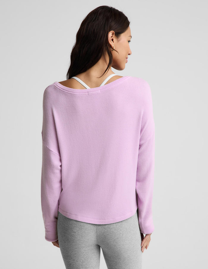 A woman shown from the back wearing a light purple long-sleeve knit top with a wide, open back design that reveals white underwire bra straps, paired with gray leggings, against a light gray studio background.