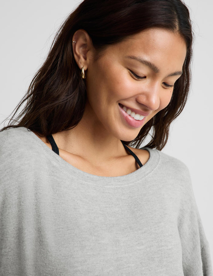 A close-up of a woman wearing a light gray crewneck sweater, showing her smiling and looking down. She has dark brown hair and is wearing a small gold hoop earring.