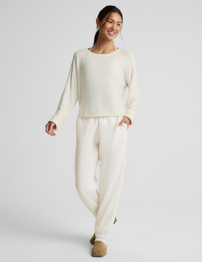 A woman modeling a matching cream lounge set consisting of a long-sleeve crewneck top and relaxed-fit pants, paired with tan slip-on shoes against a light gray studio background.