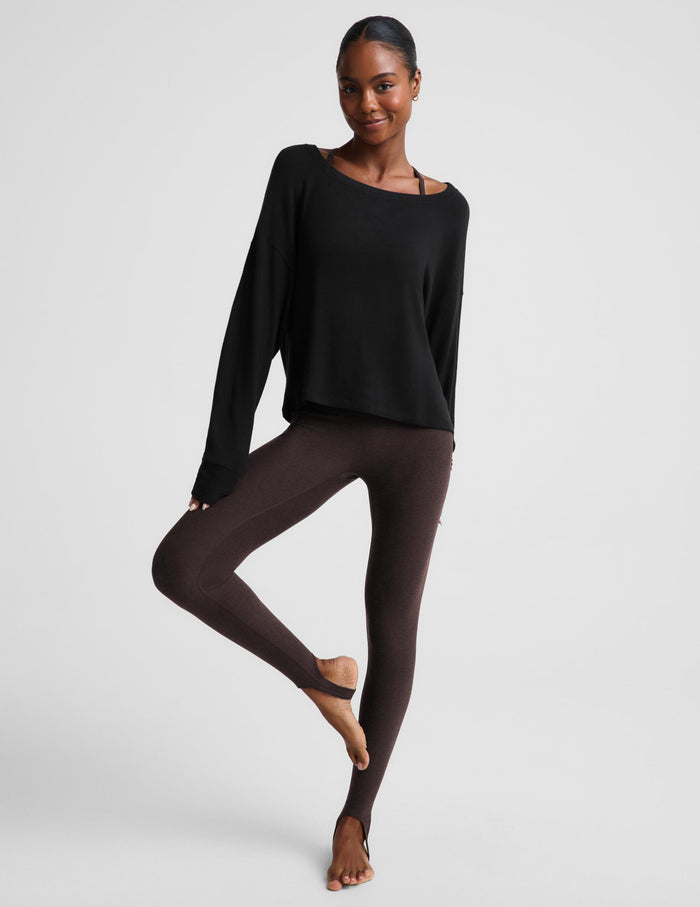A woman modeling a black long-sleeve scoop-neck top with a relaxed fit, paired with dark brown leggings, against a solid light gray background.