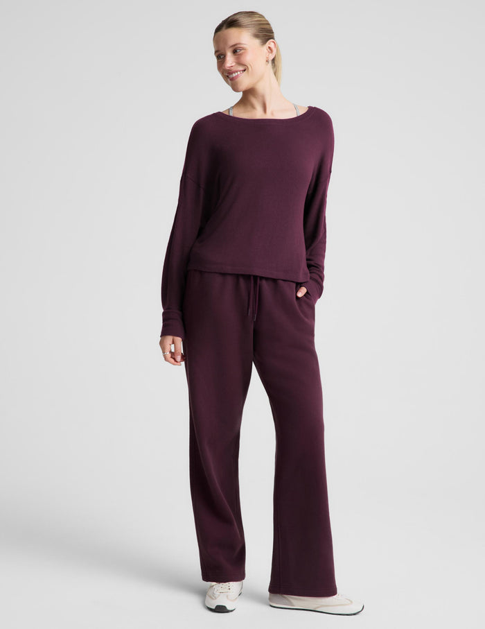 A woman modeling a matching deep plum purple loungewear set, featuring a long-sleeve boat-neck top and coordinating sweatpants, against a solid light gray background.