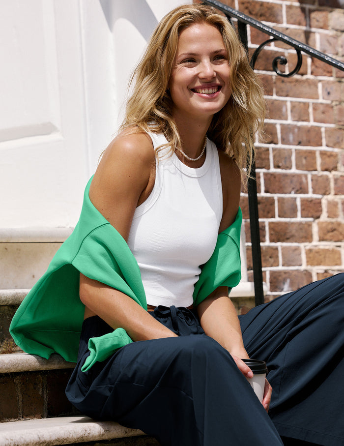 A woman sitting on outdoor brick steps wearing a white ribbed tank top, dark navy wide-leg pants, and a bright green shrug draped off her shoulders.