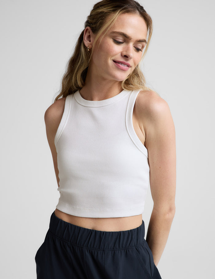 Woman wearing a white ribbed cropped tank and blue pants against a plain background.