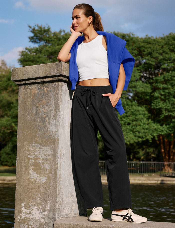 A woman stands outdoors by a stone pillar next to water, wearing a white ribbed crop top, black wide-leg drawstring pants, a bright blue open jacket, and white sneakers.
