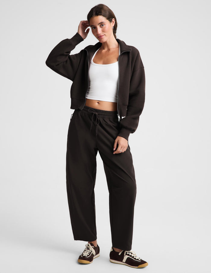 A woman modeling a dark brown zip-up hoodie, matching dark brown sweatpants, a white ribbed tank top, and brown sneakers with white soles, posed against a light gray studio background.