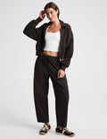A woman modeling a dark brown zip-up hoodie, matching dark brown sweatpants, a white ribbed tank top, and brown sneakers with white soles, posed against a light gray studio background.