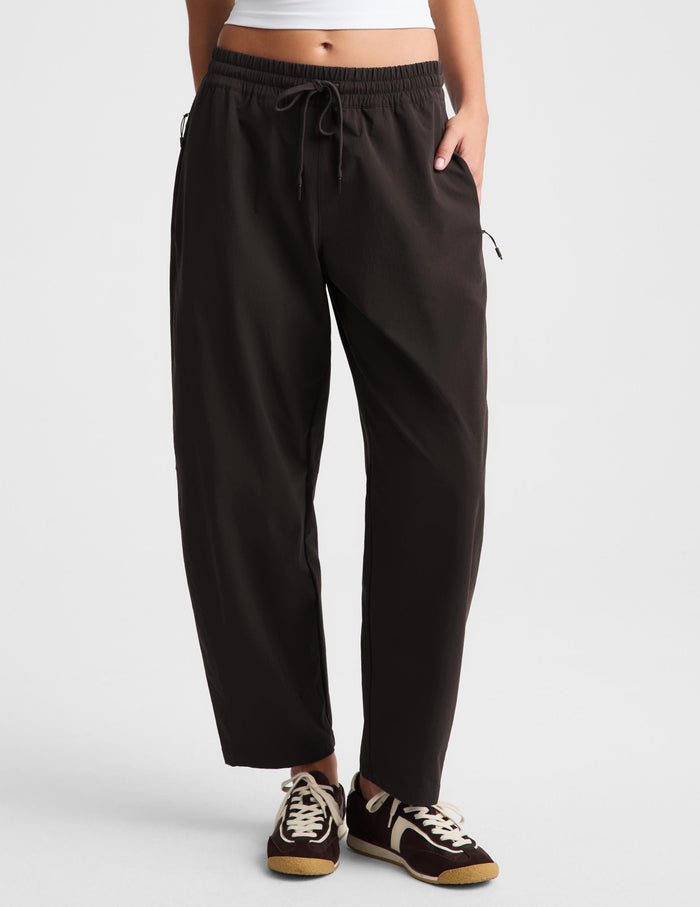 A person wearing loose brown drawstring pants with side pockets and black sneakers, shown from the waist down against a light gray studio background.