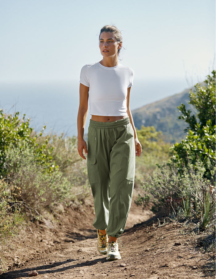 Woman walking on a trail wearing the olive green Peak Pant with cargo pockets and a gathered elastic waistband.