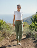 Woman walking on a trail wearing the olive green Peak Pant with cargo pockets and a gathered elastic waistband.