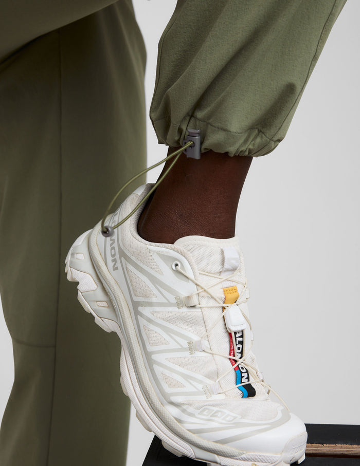 Detail of the olive Peak Pant featuring an adjustable bungee hem toggle cinched above a white hiking shoe.