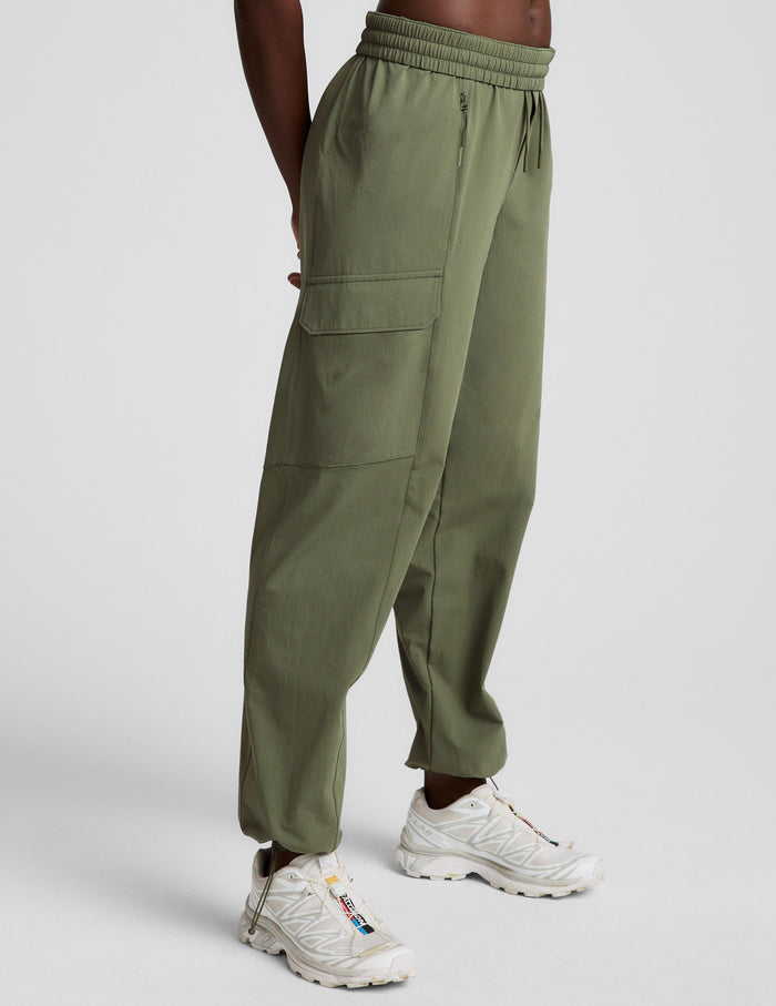 Side view of olive green Peak Pant cargo joggers with an elastic waist, patch pocket, and bungee hem with white sneakers.