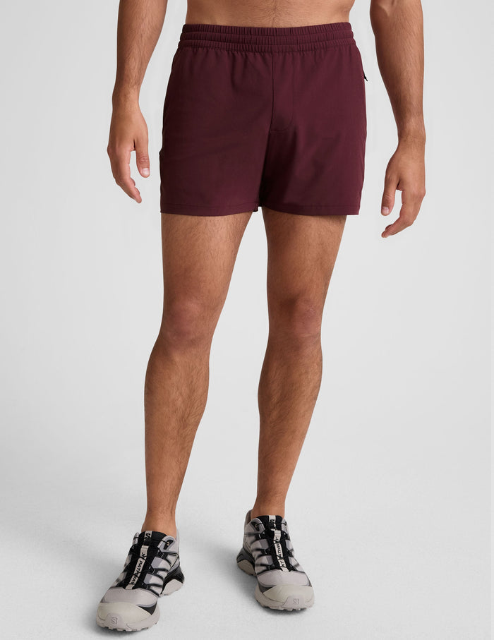 A man wears Pivotal 2.0 Men's Short 5" in maroon with an elastic waistband, styled with athletic shoes on a white background.