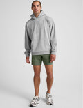 Man wearing olive Pivotal 2.0 Men's Short 7" with a grey hoodie, showing the relaxed mid-thigh fit and elastic waist.