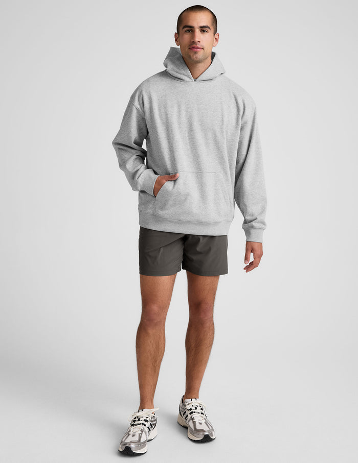 A man wears dark gray Pivotal 2.0 Men's Short 7" with a light gray hoodie and white sneakers against a white background.