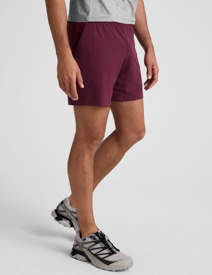 A man wears Pivotal 2.0 Men's Short 7" in maroon with an elastic waist, side pockets, and grey running shoes.