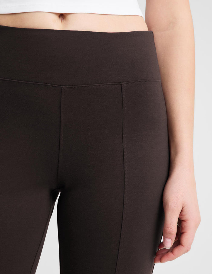 Close-up of a model wearing high-waisted dark brown leggings, showing the waistband, front seam, and hand resting by the leg.