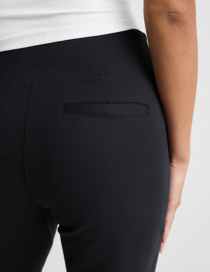 Close-up back view of a person wearing black high-waisted pants with a side welt pocket, paired with a white top, against a light gray studio background.