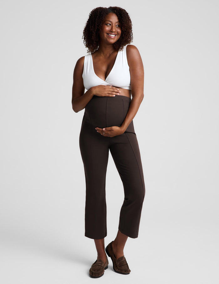A smiling pregnant woman stands against a light gray studio background wearing dark brown maternity pants and a white bra, with her hands resting on her belly.
