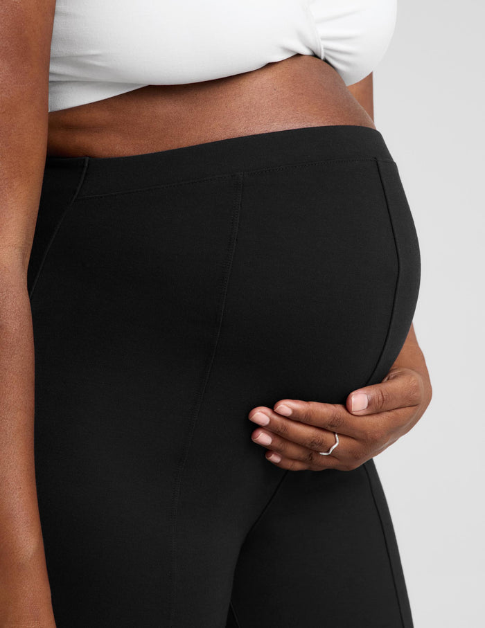 Close-up of a person wearing black maternity pants and a white bra, holding their baby bump against a light gray studio background.